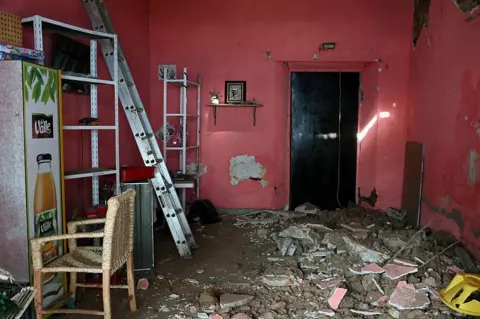 An image showing damage inside a pink house after a 6.5 magnitude earthquake in the community of San Marcos, Guerrero state, Mexico. The floor is covered in a pile broken concrete, rubble, and dirt, while a ladder is positioned on the left near shelves and a chair.