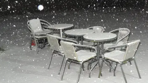 Practical Bill/WEATHERWATCHERS Garden furniture covered in snow. There are three round tables in a row, surrounded by seven chairs. Two of the chairs are stacked on each other. Snow is falling.