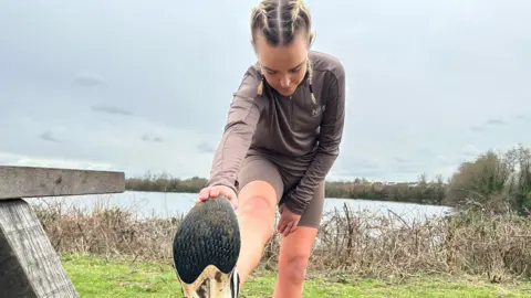 A woman wearing brown running gear stretches her right leg ahead of a training session. She stands in front of a lake, surrounded by vegetation.