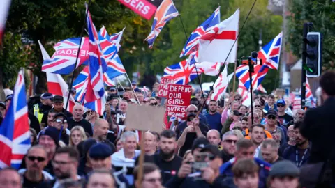 A large crowd marches down a road in Epping during a protest. Numerous Union flags and several St George’s Cross flags are prominently displayed, held high on poles throughout the group. Police officers in hi-vis jackets are along the edges of the crowd. The setting appears to be a street lined with trees.