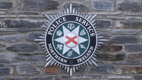 The PSNI logo on a grey brick wall. 