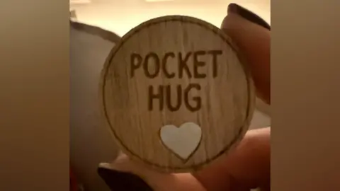 Rebecca Evans A small wooden token being held between a person's finger and thumb. On it the words pocket hug as well as a small heart can be seen. 