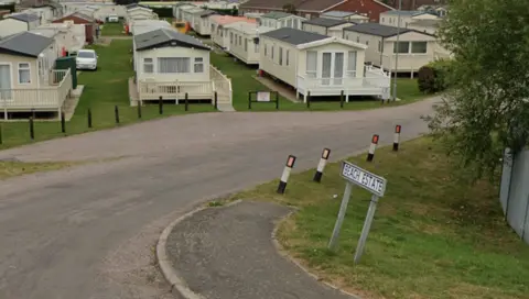 Google view of rows of static caravans. There is a Beach Estate road sign in the foreground and a number of bollards along the side of the road.