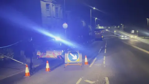 A police car with its blue lights on at the scene of a cordon along a busy road in Plymouth at night. A police officer is stood next to the car. An ambulance and a second police car are parked up further down the road. Two cars with their lights on are driving on the opposite side of the road. Traffic cones and a sign telling motorists to move to the right are next to the police car in the foreground.