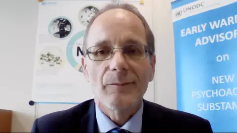 Martin Reiselhuber sits in his office, facing the camera wearing a navy blue blazer, pastel blue shirt, navy blue striped tie and glasses. Behind him are two unfurled banners. The one to his left refers to the early warning advisory message on new psychoactive substances (NPS) from the United Nations Office on Drugs and Crime (UNODC). Above his right shoulder is a second banner explaining the effectiveness of NPS.