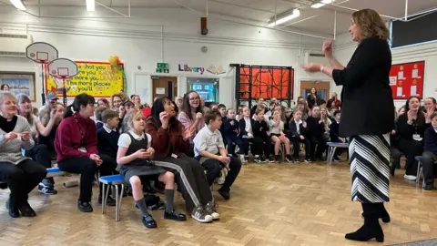 A teacher is leading an assembly in front of a group of pupils at Field Lane primary school.