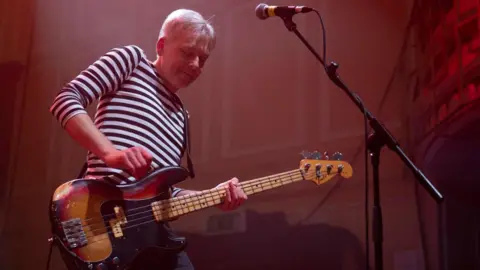 Michael Matthews Rob Tickell is performing on stage with an electric guitar. He is wearing a striped shirt and smiling.