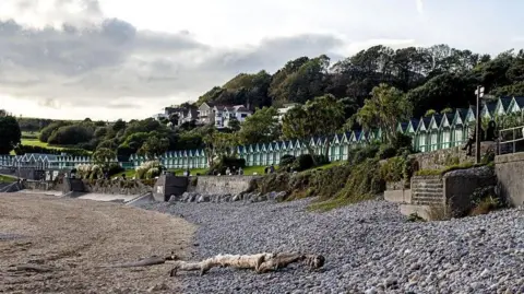 Getty Images A picture of the beach huts in Langland Bay taken from the beach. The huts curve around the edge of the beach above stretches of wall and a grassy slope underneath the huts. There is sand visible on the left of the picture and pebbles on the right. A large washed up log sits on top of the pebbles. A few people wearing jackets or long-sleeved clothes can be seen walking on the promenade between the beach and the grassy slope. Behind the huts are trees with a number of houses among them in the centre of the picture. The sky is cloudy but bright, reflecting the fact that the photo is taken in October and is not from the same day as the rest of the photos in the article.