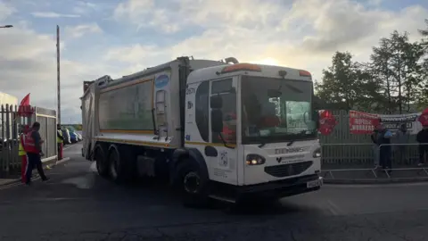 A large white bin lorry driving out of the exit of a depot. There are large metal fences surrounding it, with red unite the union flags on them.