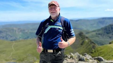 Leslie Baron with his thumbs up on a mountainside 