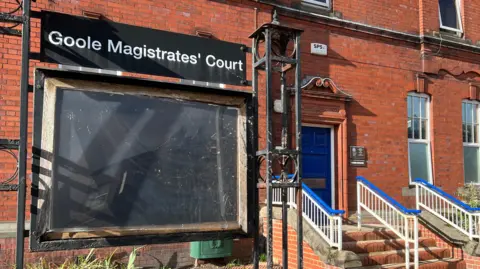 A black noticeboard with a sign reading: 'Goole Magistrates' Court' above it. The board is in front of a redbrick building and on the right is a cobalt blue door with a row of three steps leading up to it