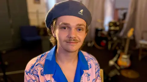 Martin Giles/BBC Reece Pasby sits in front of an out-of-focus drum kit and guitar. He wears a flat cap and an open-collar Cuban shirt. The shirt features a bright blue and pastel pink design.