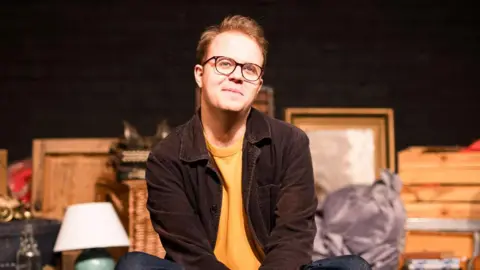 Man wearing black-rimmed glasses smiling while sat on crossed-legged on a theatre stage