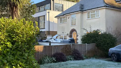 A person in an all white forensic costume covering their whole body can be see walking towards the front doo of a house which has a boat on the driveway