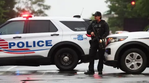 A police officer with a gun stands in front of two white marked police cars
