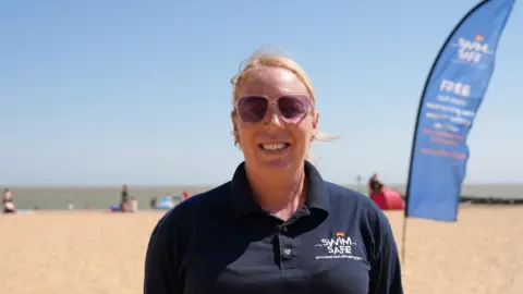 Qays Najm/BBC Allyce Clarke of Swim Safe stands on a sandy beach. She is wearing a navy blue polo shirt and pink sunglasses. She has blonde hair and is smiling into the camera. Behind her people are playing on the beach.