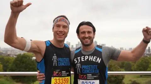 Two men in black and blue running vests with their arms in the air, one of them with a white patterned headband