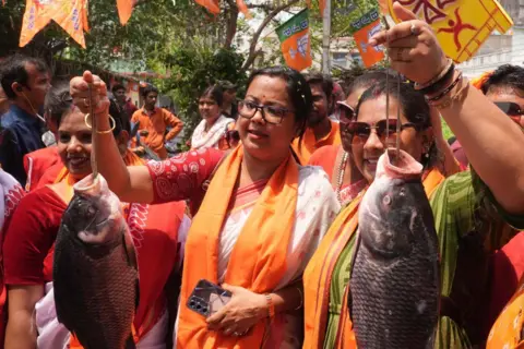 Women politicians wearing red-and-white saris and saffron scarves around their necks hold up fish