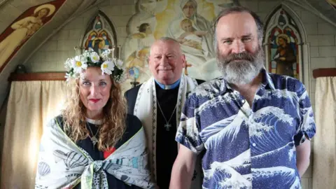 Julie Wilson Nimmo and Greg Hemphill stand in front of the Rev Chris Wallace at the Italian Chapel in Orkney. The chapel has a religious mural on tiles with curtains in the background. Julie wears flowers in her hair with a shawl tied round her and Greg wears a short sleeve, blue shirt with wave illustrations.