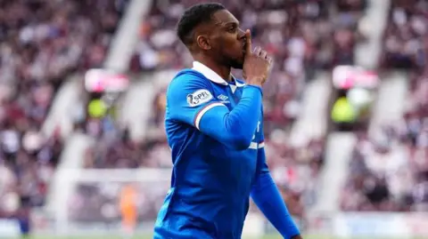 Rangers footballer Dujon Sterling wearing a blue football strip. He has his hand in front of his mouth, representing a shush sign. Behind him are blurred-out football fans in a stand.