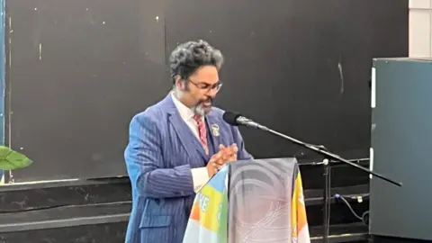 Bishop Dr Desmond Jaddoo a man in a blue pinstripe suite with a red tie is talking into a microphone