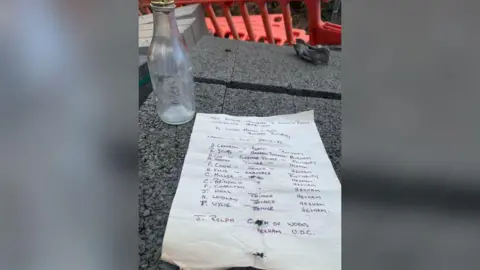 A clear glass bottle on a table alongside a handwritten document listing several people's names, job titles and places of origin. 