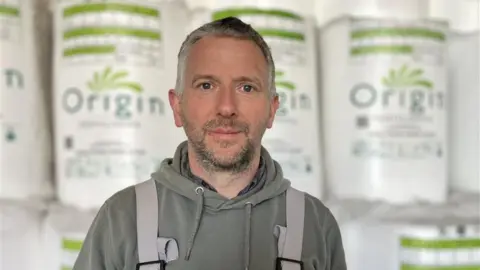 A man in farmers braces stands in front of tubs of fertiliser. He has short grey hair and stubble.