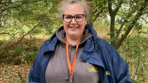 A woman with grey hair pulled back in a ponytail is smiling at the camera. She is wearing a navy jacket and grey hooded top, with an orange lanyard around her neck. In the background is healthy woodland