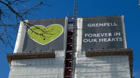 A black and white sign covers Grenfell Tower which reads 'Grenfell, Forever in Our Hearts', with a clear blue sky in the background. 