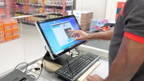 G4S A hand touching a touchscreen till inside a supermarket. The shopworker is wearing a black and red short sleeve t-shirt.