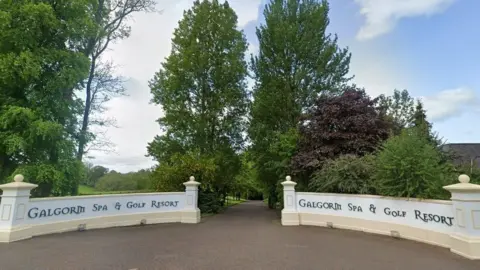 Google Google view of the Galgorm spa and golf resort, a white curved wall with text, with a brown driveway and large green trees