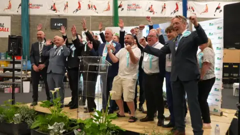 Lots of men in suits with blue rosettes, and one woman, stand on a stage cheering with their arms raised in triumph. 