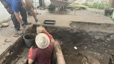 Lara King/ BBC An archaeologist wearing a red polo shirt and a straw hat is standing in a trench. There is a man wearing a blue polo shirt and flat cap who is passing a white container and a black bucket to the person in the trench. A wheelbarrow and a wooden handle (possibly a spade) are also visible. 