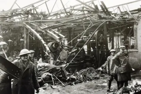 Trevor Frecknall Black and white photo of men in steel hats and cloth caps inside a badly damaged factory