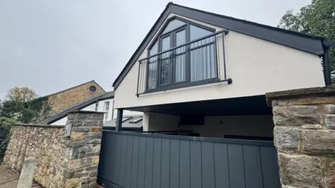 The outside of the house which has a cream-painted top floor with grey framed windows and a gate over them and a grey fence below