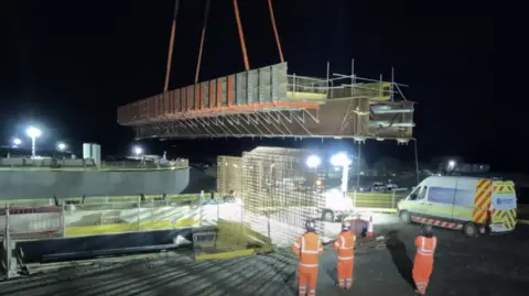 A large long structure suspended on a crane as three engineers look on