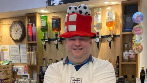 BBC / Ian Wood Sean Rowbottom is standing behind the bar and smiling brightly at the camera. He is wearing a tall red hat with an England flag on it and three fluffy football balls on top. He has stubble and is wearing a white England kit.
