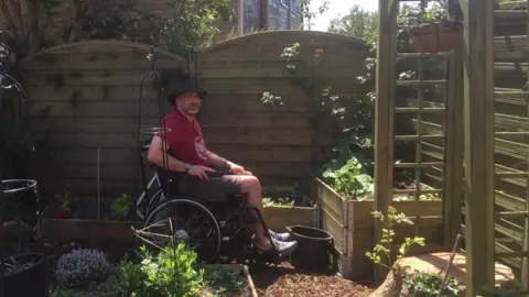 Julie Heath A man in a manual wheelchair on a sunny day between wooden raised beds in a garden and in front of wooden fence. He is wearing a hat and looking towards the camera