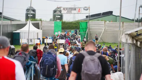 Ben Birchall/PA Media A large crowd of people leaving Glastonbury. They are all heading towards pedestrian gate A. They are all carrying luggage and camping equipment.