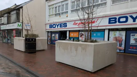 LDRS Two large stone squared plant pots on a high street. Small leafless trees grow from them. They are outside a Boyes shop and it has been raining.