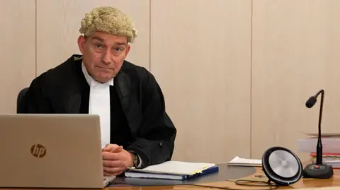 Sheriff Alastair Carmichael, a 62-year-old man, wearing a legal wig and outfit, sitting on the bench at Dundee Sheriff Court. He has a laptop in front of him. There is a microphone and speaker on the bench.