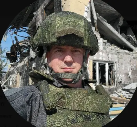 X (Twitter) A man wearing military gear in front of a bombed-out building