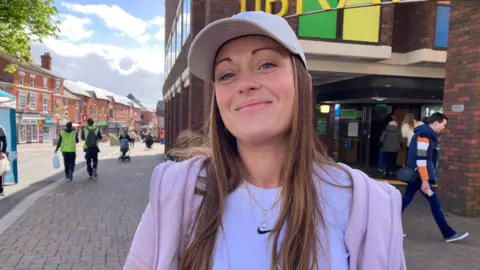 A woman wearing a white top, pink hoodie and a white cap, standing in front of Redditch Library.