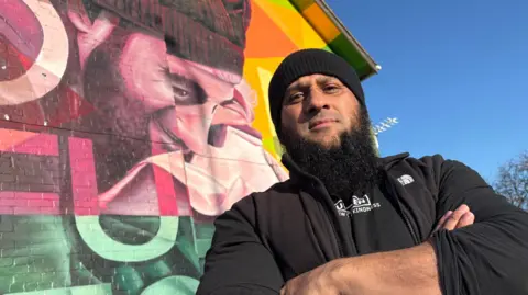BEN SCHOFIELD/BBC Atiq Rehman standing in front of a brightly coloured mural outside. The shot is taken from below at a slightly skewed angle, looking up and to the left of frame. He is on the right of frame, wearing a black tight fitted knitted hat, black fleece jacket, which is unzipped, and a black top. He has a long black beard and is looking directly down the camera. The mural behind him shows a man – who is also Atiq – smiling and leaning forward, though he is distorted by the shape of the building. There are also letters that form another part of the mural. The sky in the top right corner is bright blue and cloudless. 