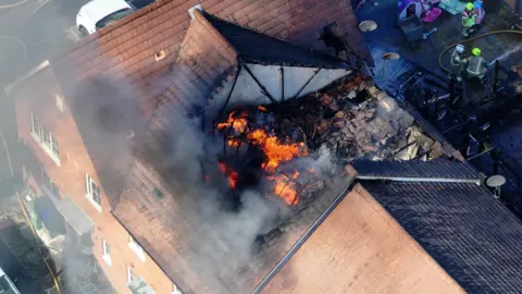 Eddie Mitchell An aerial view of a house on fire with flames seen in the roof and firefighters in the garden behind the house.