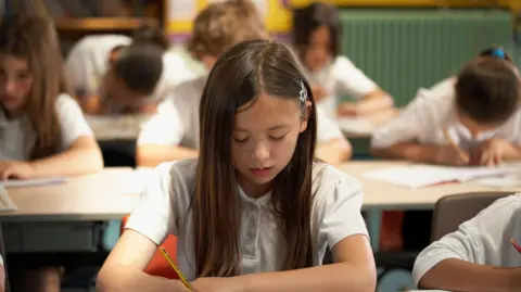 A young girl with long-brown hair holds a yellow and black pencil in her hand. She wears a white polo top. Behind her are five young people in the same outfit, holding similar pencils writing on white pages. 