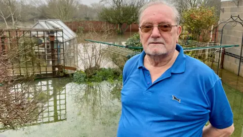 A man in a blue polo shirt and dark glasses is standing in a flooded domestic garden. Bushes, plants, a garage, greenhouse and washing pole is partially submerged. There is a wood panelled fence around the garden. Trees stand behind the fence in a field which is also flooded.