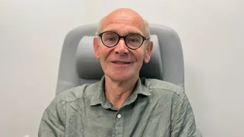 A man wearing round glasses and a light green shirt sits in a high-backed chair against a plain wall in a clinical setting.”