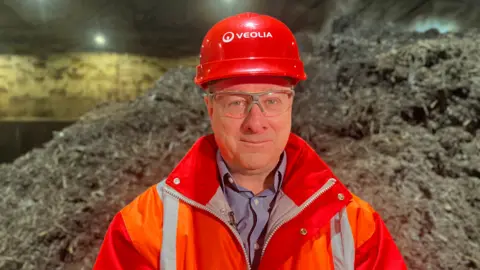 Lucinda Adam/BBC A man is standing facing the camera wearing an orange hi-vis jacket and hat, with Veolia written on the hat in white