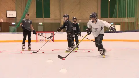 Four people playing roller hockey in a big hall. They are holding large hockey sticks and wearing oversized black and white jerseys and helmets with face guards.